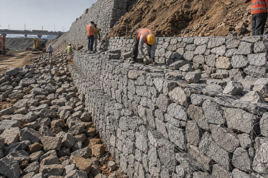 Stone retaining wall construction in progress near a bridge embankment